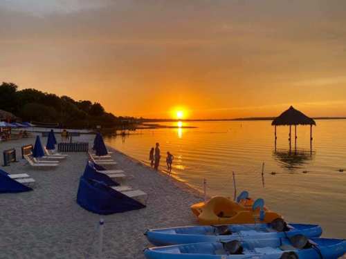 Sunset over a calm beach with silhouettes of people walking, kayaks on the shore, and a thatched hut in the distance.