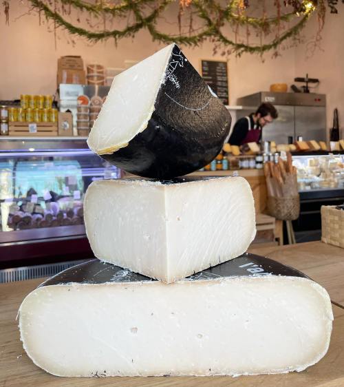 A stack of three cheese wheels in a shop, with a blurred background of a counter and a person working.