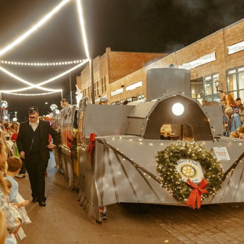 A festive train float decorated with lights and wreaths, parading down a street lined with spectators at night.