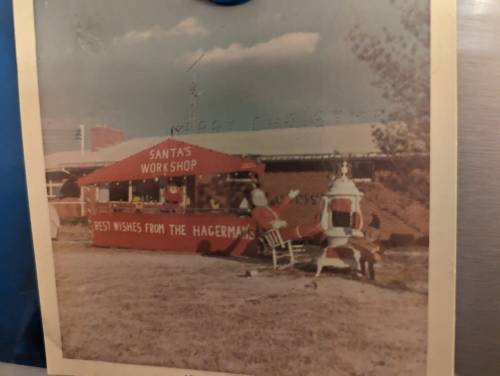 A vintage photo of Santa's Workshop with a red building and festive decorations, set in a grassy area.
