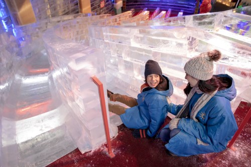 Two children in winter coats sit on an ice slide, smiling and enjoying the colorful ice sculptures around them.