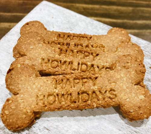 Three bone-shaped dog treats with "Happy Howlidays" engraved on them, arranged on a wooden surface.