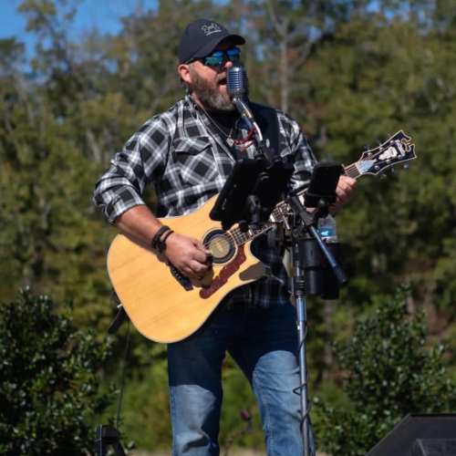 A man in a plaid shirt and cap sings into a microphone while playing an acoustic guitar outdoors.