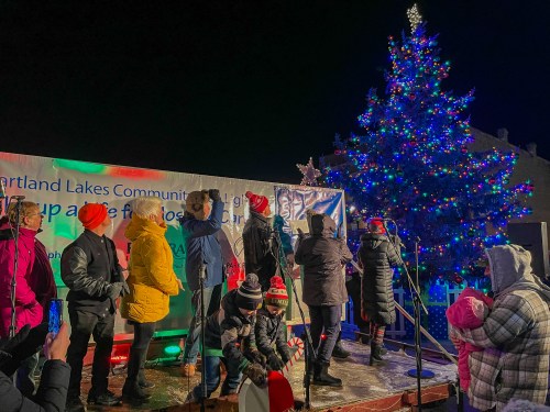 A festive gathering with people in winter attire celebrating by a brightly lit Christmas tree at night.