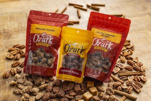 Three bags of Ozark nut roaster pecans—praline, cinnamon, and salted caramel—surrounded by pecans and caramel pieces.