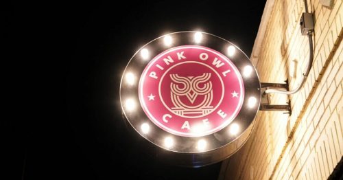 Neon sign for "Pink Owl Cafe" featuring an owl graphic, illuminated against a brick wall at night.