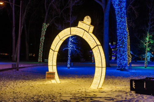A large illuminated ring-shaped decoration in a snowy park, surrounded by colorful lights on trees at night.