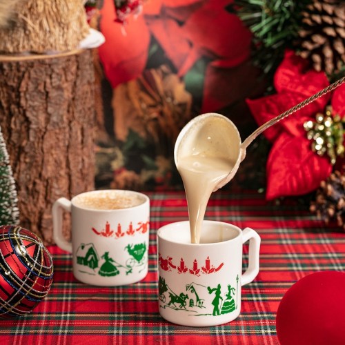 A festive scene with two mugs, one being filled with creamy drink, surrounded by holiday decorations and a plaid tablecloth.