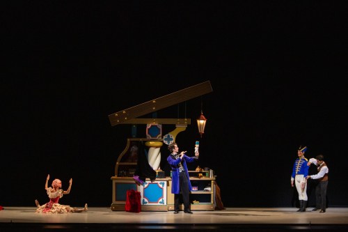 A theatrical scene with performers in costumes, featuring a man holding a lantern and a whimsical set design.