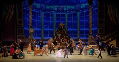 A festive ballet scene with dancers in colorful costumes performing around a large Christmas tree on stage.