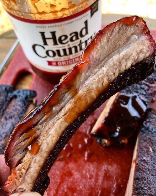 Close-up of a juicy, smoked rib with a rich glaze, held above a bottle of Head Country barbecue sauce.