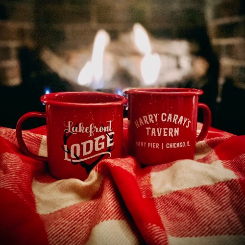 Two red mugs with logos, resting on a checkered blanket in front of a cozy fireplace.