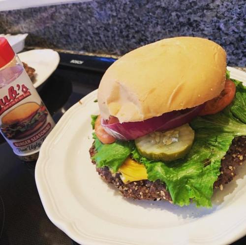 A large burger with lettuce, tomato, pickles, and onion on a plate, next to a bottle of burger seasoning.