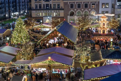 A festive holiday market with decorated stalls, twinkling lights, and a large Christmas tree, bustling with visitors.