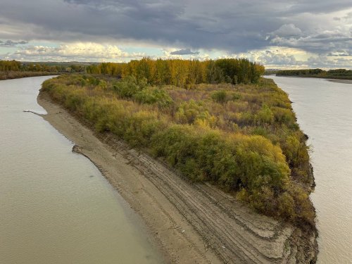 A river bends around a lush, green island with autumn-colored trees under a cloudy sky.