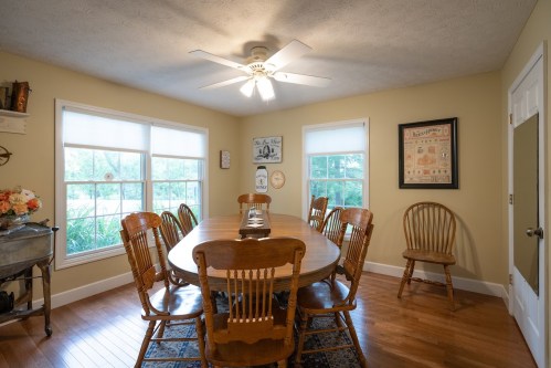 A cozy dining room with a large wooden table, eight chairs, a ceiling fan, and windows letting in natural light.