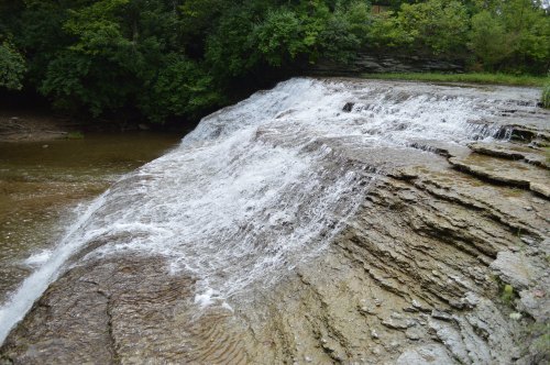 A serene waterfall cascading over rocky ledges into a calm stream, surrounded by lush green trees.