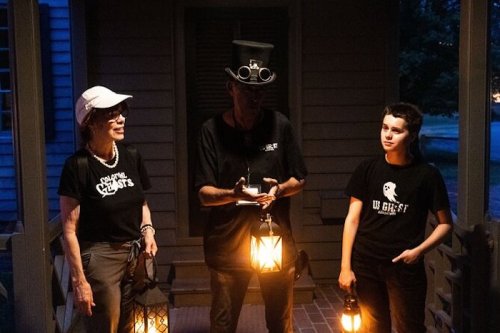 Three people stand on a porch at night, holding lanterns, wearing black shirts, with one in a top hat.