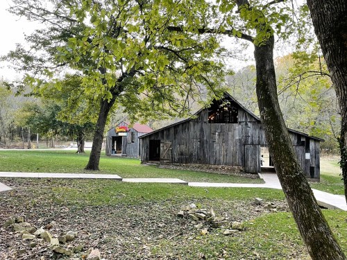 A rustic wooden barn surrounded by trees and a grassy area, with a pathway leading through the landscape.