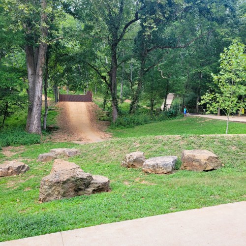 A grassy area with rocks, trees, and a dirt path leading to a small hill in a park setting.
