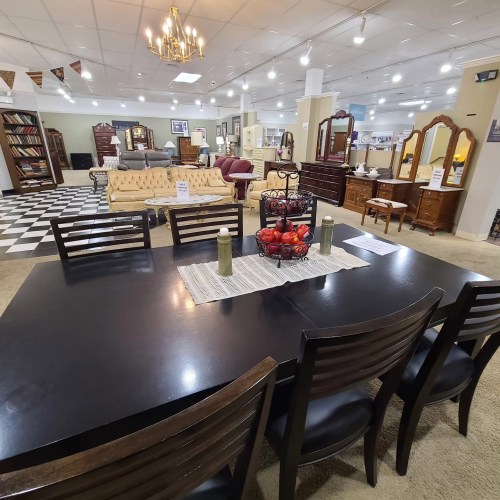 A spacious furniture showroom featuring a dining table, chairs, and various sofas and dressers in the background.