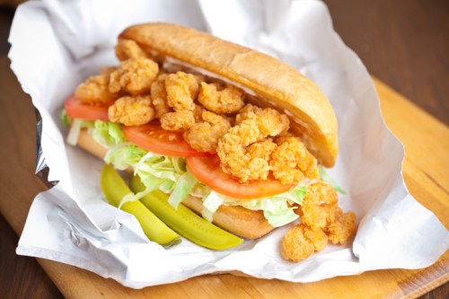 A sandwich filled with crispy shrimp, lettuce, and tomato, served in a soft bun with pickles on the side.