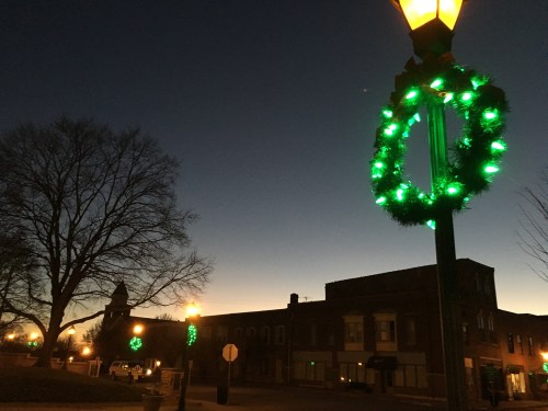 A streetlamp adorned with green wreaths against a twilight sky, with buildings and trees in the background.