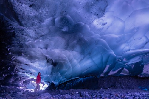 A person in a red jacket stands inside a stunning ice cave, illuminated by soft blue and purple light.