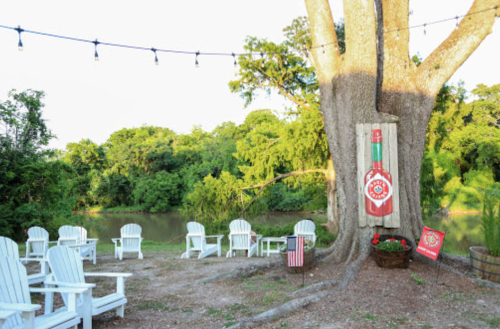 A serene outdoor space by a river, featuring white chairs, a large tree, and decorative lights.