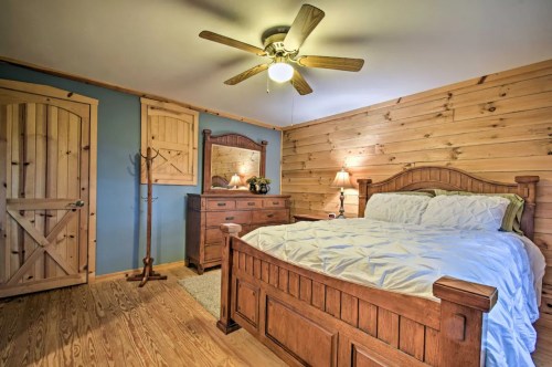 Cozy bedroom with wooden walls, a ceiling fan, a bed, dresser, and mirror, featuring warm lighting and rustic decor.