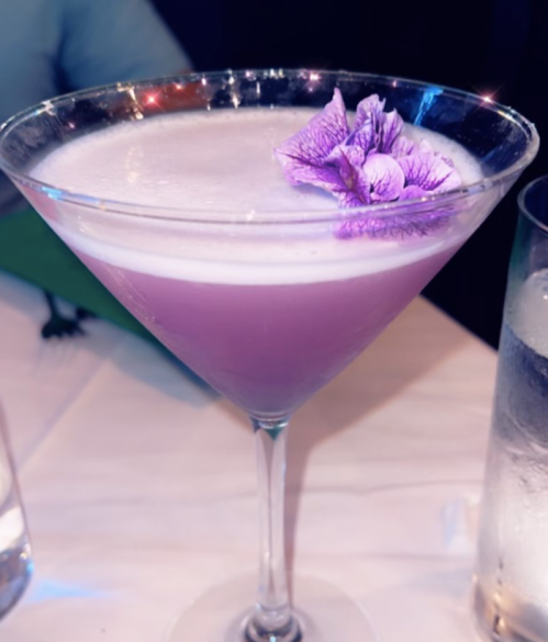 A pink cocktail in a martini glass, topped with a purple flower, sits on a table with a glass of water nearby.