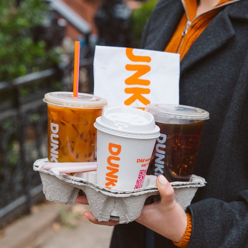 A person holds a tray with three Dunkin' drinks and a bag, set against a blurred outdoor background.