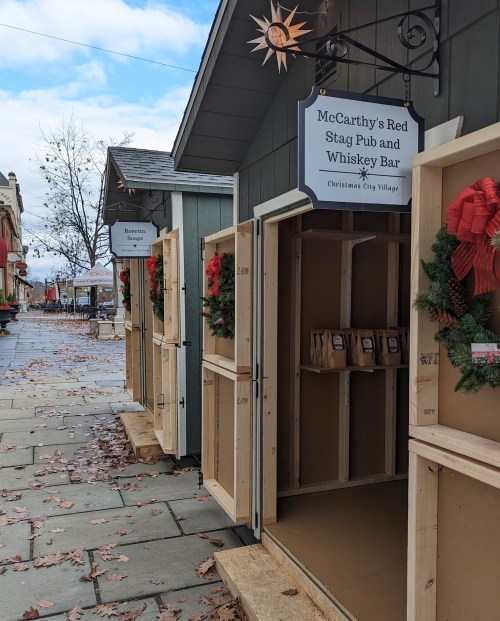 Wooden stalls with festive wreaths, labeled for McCarthy's Red Stag Pub and other shops, in a quaint village setting.