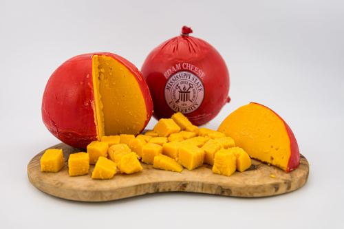A round red cheese wheel with a cut section, surrounded by diced yellow cheese on a wooden board.