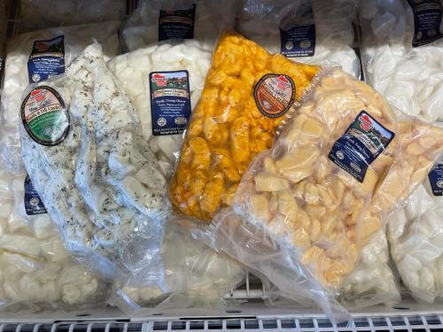 Packages of fresh cheese curds in various flavors, including white, orange, and herb-infused, displayed in a freezer.