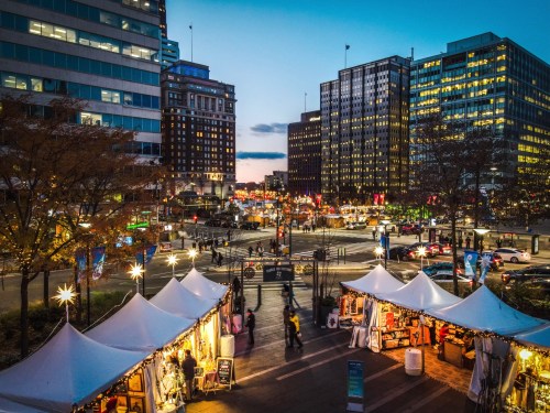 A bustling city street at dusk, featuring market stalls and illuminated buildings in the background.