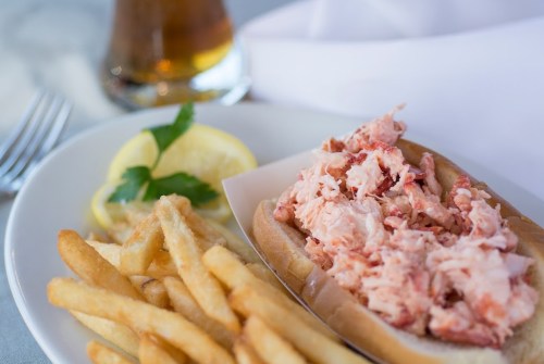 A plate with a lobster roll, fries, lemon slices, and a drink in the background.
