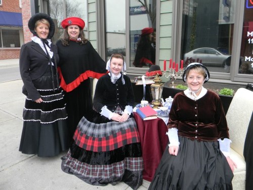 Four women in vintage dresses pose together outside a building, with a table set for tea and decorative items.