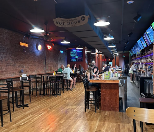 A lively bar with wooden floors, brick walls, and patrons enjoying drinks at the counter and tables.