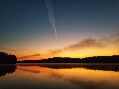 A serene lake at sunrise, reflecting vibrant orange and blue hues with a misty shoreline and a wispy cloud in the sky.