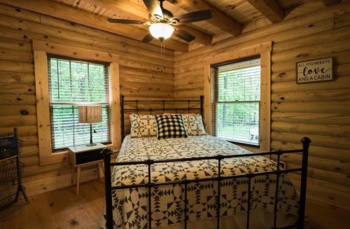 Cozy log cabin bedroom with a bed, patterned bedding, wooden walls, and two windows letting in natural light.