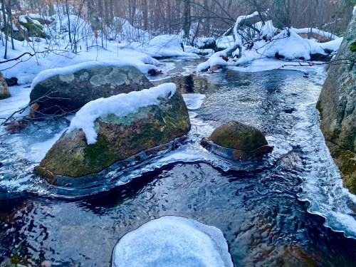 A serene winter scene featuring a stream with rocks, partially covered in snow and ice, surrounded by trees.