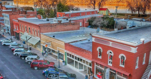 Aerial view of a small town street with brick buildings, parked cars, and a river in the background during sunset.
