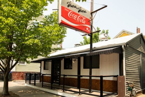 A small, one-story building with a Coca-Cola sign and a tree in front, set in a residential area.