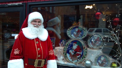 Santa Claus stands outside a shop window decorated with festive holiday plates and lights.