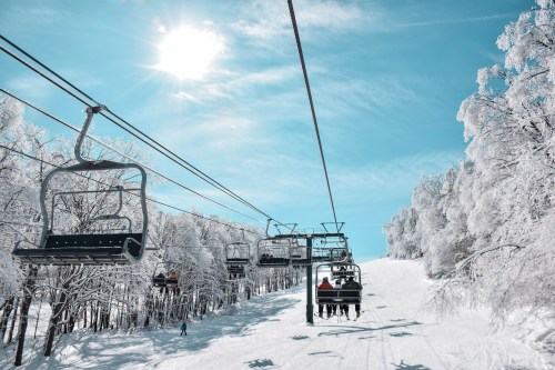 Ski lift chairs ascend a snowy slope under a bright sun, surrounded by frosted trees and a clear blue sky.
