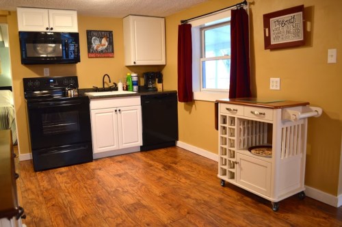 A cozy kitchen with black appliances, white cabinets, and a wooden cart on the floor, featuring warm yellow walls.