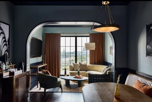 A stylish hotel room featuring a cozy seating area, large window, and modern decor in muted tones.