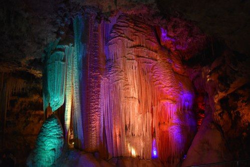Colorful stalactites and stalagmites illuminated in a cave, showcasing vibrant hues of blue, green, purple, and red.