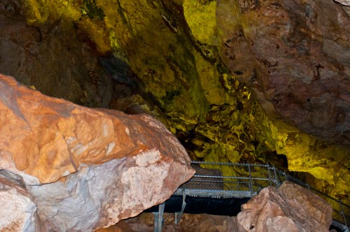 A rocky cave interior with yellow-lit walls and a metal walkway among large boulders.